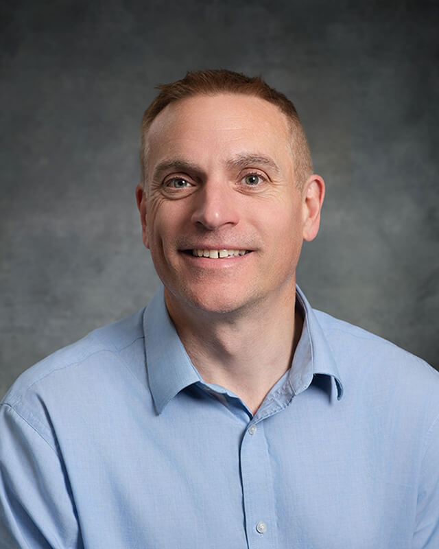 smiling headshot of company director in blue shirt against a mottled grey background